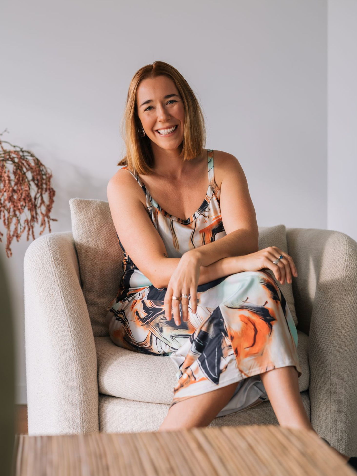 Kate Holland sitting on a couch wearing a floral dress, smiling at the camera.